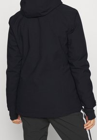 Giacca impermeabile di un blu navy scuro con una forma aderente, cappuccio attaccato e texture liscia. Presenta aperture per i pollici ai polsini delle maniche e un taglio sartoriale.