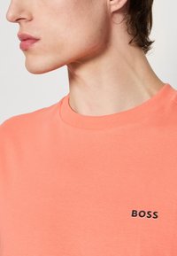 Korálové tričko s krátkým rukávem, které má žebrovaný kulatý výstřih a malé černé logo "BOSS" na hrudi. Hladká textura a jednoduchý design.