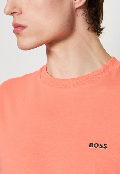 T-shirt corail à manches courtes avec un col rond côtelé et un petit logo "BOSS" noir sur la poitrine. Texture lisse et design simple.