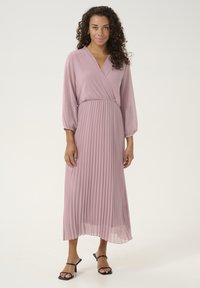 Pleated, midi-length dress in soft pink chiffon with a wrap design, v-neckline, and long sleeves; worn with black strappy sandals.