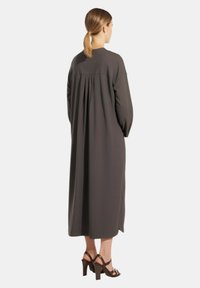 Abito lungo grigio scuro con vestibilità comoda, che presenta un dettaglio plissettato sul retro e maniche lunghe. Indossato con sandali con tacco alto marroni.