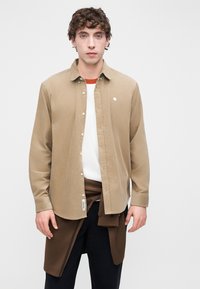 Μπεζ πουκάμισο με κουμπιά, φτιαγμένο από μαλακό ύφασμα, με λογότυπο στο στήθος, φορεμένο πάνω από λευκό πουκάμισο με καφέ περιδέσιμο στη μέση, συνδυασμένο με σκουρόχρωμο παντελόνι.