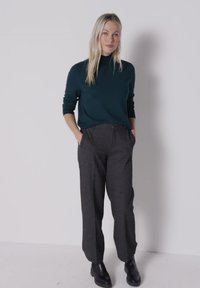 Donna con lunghi capelli biondi che indossa una maglietta verde scuro a maniche lunghe, pantaloni grigio scuro e stivaletti neri, in piedi con le mani nelle tasche.