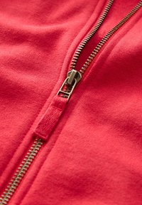 Detailní záběr částečně rozepnutého kovového zipu na červené látce, zobrazující zuby zipu a texturu látky.