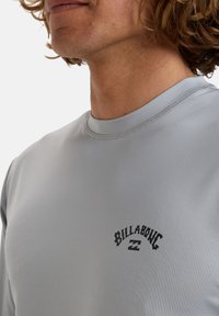 Camisa gris de manga larga hecha de una tela suave y ligera. Presenta un logo negro de "Billabong" en el pecho y un cuello redondo.