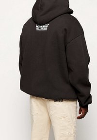 Dark gray hoodie with a large hood, loose fit, featuring a logo on the back. Paired with light beige cargo pants, highlighting pocket details.