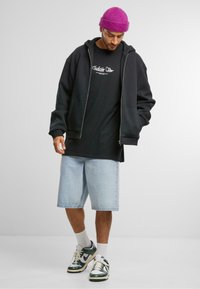 Černá zipová mikina, černé tričko, světle modré denimové kraťasy, zelené a bílé tenisky a fialová pletená čepice. Uvolněný streetwear styl.