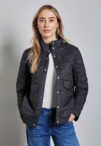 Schwarze gesteppte Jacke mit Stehkragen, vorderem Druckknopfverschluss, zwei Brusttaschen und langen Ärmeln, kombiniert mit einem weißen Oberteil und blauen Jeans.