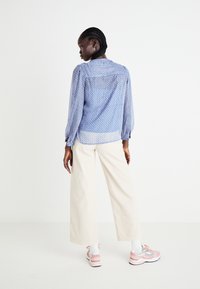 Blusa azul con estampado, mangas largas abullonadas, un brillo sutil y detalle de botones en la espalda, combinada con pantalones de pierna ancha color beige claro.
