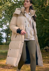 Beige pufferjakke med høj krave og trykknapper, lagdelt over en lys mønstret sweater og olivengrønne leggings, i en naturlig omgivelser.
