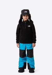 Pulover din fleece negru cu fermoar până la jumătate, pantaloni de ski albastru strălucitor și negru, și o pălărie neagră cu clapete pentru urechi din blană. Logo-urile de pe haine sunt vizibile.