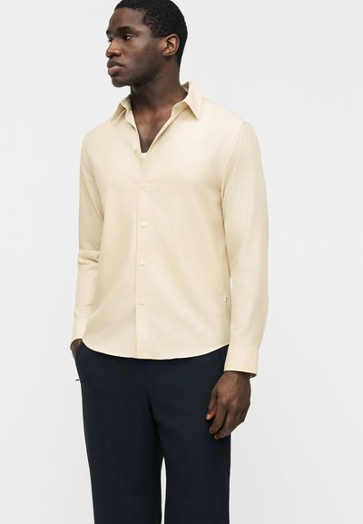 Άνδρας με μπεζ πουκάμισο με κουμπιά και μακριά μανίκια και σκούρο παντελόνι, στέκεται με τα χέρια στις τσέπες, κοιτάζοντας προς τα πλάγια.