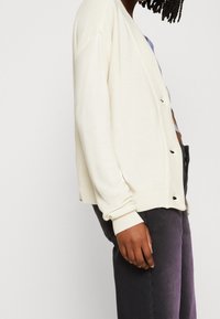 Gräddfärgad cardigan med ribbad textur, med knappar och en avslappnad passform, bärs över mörkgrå byxor.