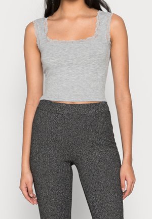 Vrouw die een mouwloze grijze geribbelde cropped top met kantafwerking draagt en hoog getailleerde, getextureerde donkere grijze leggings, staand tegen een effen achtergrond.