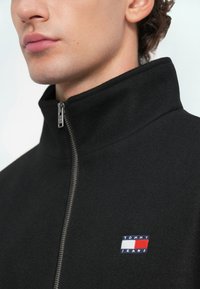 Μαύρο μπουφάν με ψηλό γιακά και φερμουάρ μπροστά, κατασκευασμένο από μαλακό υλικό. Διαθέτει μια μικρή πολύχρωμη ετικέτα λογότυπου με την ένδειξη "TOMMY JEANS" στο στήθος.