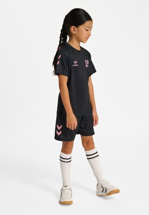 Joven chica con camiseta deportiva negra y pantalones cortos con detalles rosas, lleva calcetines blancos hasta la rodilla y zapatillas blancas, de pie sobre un fondo liso.