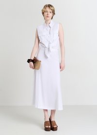 Camicia bianca senza maniche con un nodo sul davanti, abbinata a una lunga gonna bianca. Sandali platform marroni e una borsa intrecciata completano il look.