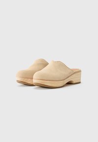 Beige mockasklädda klackar med rund tå, slip-on design, träsula och synlig söm längs kanten. Låg klack för stabilitet.