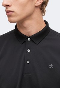 Jeune homme portant un polo noir à trois boutons avec un petit logo "ck" sur le côté gauche de la poitrine, légèrement tourné vers la gauche.
