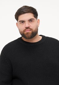 Czarny sweter robiony na drutach z fakturowanym wzorem i szeroką okrągłą szyją. Model ma krótkie włosy i pełną brodę, stoi przodem na jednolitym tle.