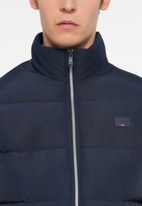 Man wearing a navy blue puffer jacket with a high collar and a visible silver zipper, featuring a small rectangular logo patch on chest.