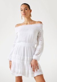 Abito bianco off-the-shoulder con maniche lunghe in pizzo, vita arricciata e gonna a strati con dettagli in pizzo.