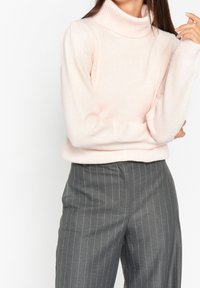 Un pull rose clair à col roulé avec un col côtelé et une texture douce, associé à un pantalon gris à rayures. Le pull présente des manches légèrement bouffantes.