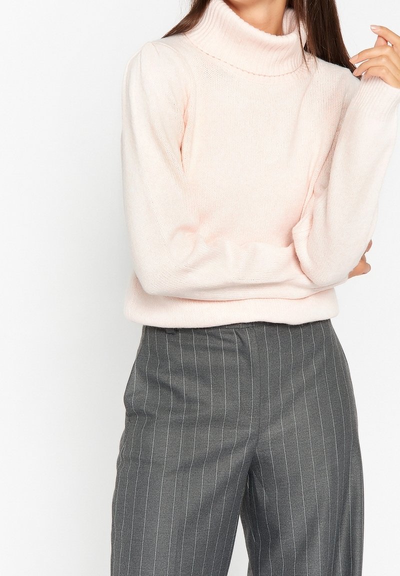 Un pull rose clair à col roulé avec un col côtelé et une texture douce, associé à un pantalon gris à rayures. Le pull présente des manches légèrement bouffantes.