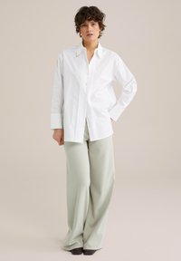 Biela košeľa na gombíky s vertikálnymi záhybmi, v kombinácii s voľnými svetlozelenými nohavicami a tmavými lodičkami. Hladká textúra látky.
