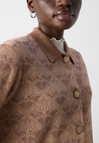 Braune strukturierte Jacke mit geometrischem Muster, mit einem Kragen und zwei runden, hellbraunen Knöpfen. Dezente schimmernde Details.