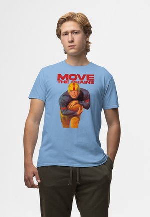 Junger Mann trägt ein blaues Hemd mit einer Illustration eines Vintage-Fußballspielers und rotem Text "MOVE THE CHAINS" auf der Brust.