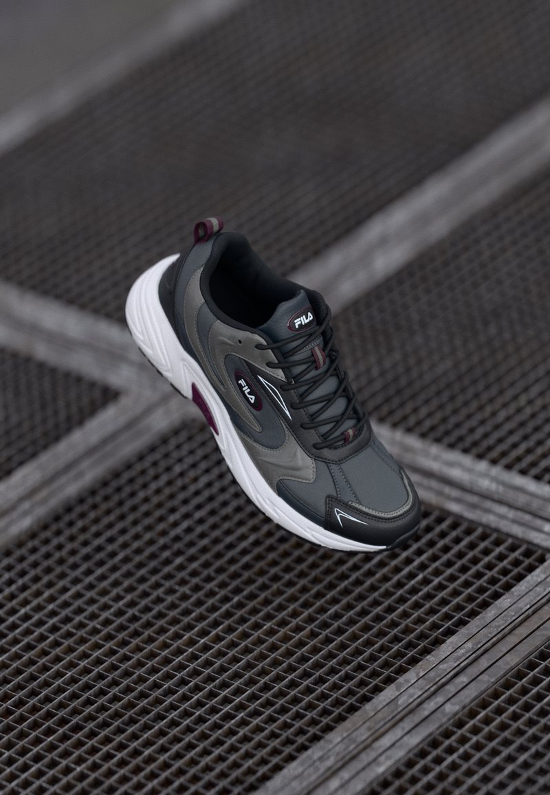 Szare buty sportowe z czarnymi i białymi akcentami, z materiału siatkowego, zaokrąglonym noskiem, wyściełanym kołnierzem oraz detalem z logo na języku i boku.
