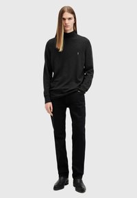Maglione grigio scuro con collo alto, realizzato in un materiale morbido. Abbinato a pantaloni neri e scarpe, in piedi davanti a uno sfondo neutro.