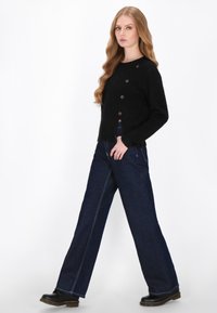 Sort strikket cardigan med rund Hals og tre store knapper, parret med vidde bukser af mørkeblå denim med hvidt syning.