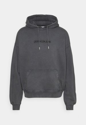 Szara bluza z kapturem, z kieszenią kangurką, ribbowymi mankietami i dołem. Posiada czarny napis "CALVIN KLEIN JEANS" na piersi.