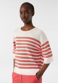 Gestreifter Pullover aus weichem Strickstoff; mit abwechselnd korallen- und weißen Streifen, weitem Ausschnitt und lockeren Ärmeln. Kombiniert mit rosa Hose.