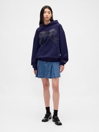 Haut à capuche bleu marine avec un grand motif de nœud accentué par des détails cloutés, associé à une jupe plissée en denim et des chaussures noires.