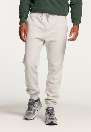 Mand iført lysegrå sweatpants, grå og hvide sneakers og en mørkegrøn sweatshirt, der står imod en hvid baggrund.