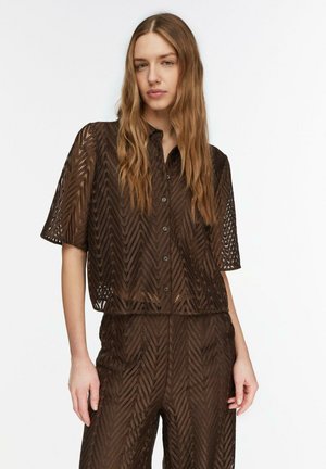 Junge Frau mit langen Haaren, die ein dunkelbraunes Spitzenoberteil mit kurzen Ärmeln und Knöpfen sowie passende, hoch taillierte Hosen mit Chevron-Muster trägt.