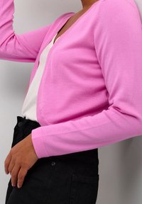 Světle růžový cardigan vyrobený z měkkého pleteného materiálu, s výstřihem do V, dlouhými rukávy a přiléhavým střihem, nošený přes bílý top.