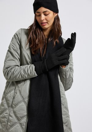 Guanti a maglia neri, sciarpa nera e una giacca trapuntata grigia chiara. La sciarpa ha una texture a coste; i guanti hanno un design senza dita con polsini a coste.