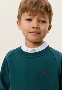 Πράσινο βαμβακερό φούτερ με rib ραφή στο λαιμό και κόκκινη κεντητή επιγραφή "II" στο στήθος, φορεμένο πάνω από λευκό πουκάμισο με γιακά.