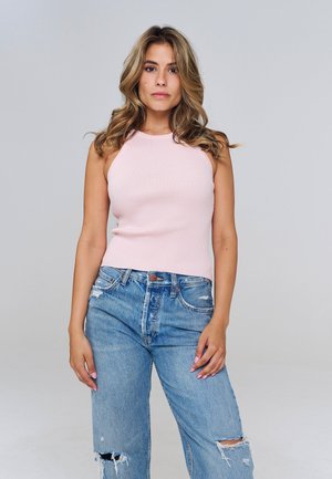 Jeune femme aux cheveux blonds ondulés, portant un top sans manches rose pâle côtelé et un jean bleu déchiré, debout devant un fond gris uni.