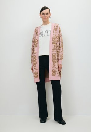 Vrouw met een roze gebloemde cardigan over een wit bedrukte blouse, zwarte broek en zwarte schoenen, staand voor een effen witte achtergrond.