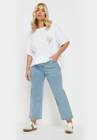 Camiseta blanca de gran tamaño con un diseño floral colorido en el pecho. Combinada con unos vaqueros rectos de color azul claro y zapatillas blancas con rayas negras.