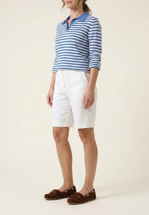 Kvinde iført en blå- og hvidstribet langærmet polo, hvide knælange shorts og brune mokkasinsko, stående sidelæns.