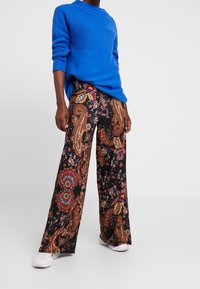 Blauwe gebreide trui gecombineerd met zwarte wijde broek met een kleurrijk paisley- en bloemenpatroon. Witte sneakers aan de voeten.