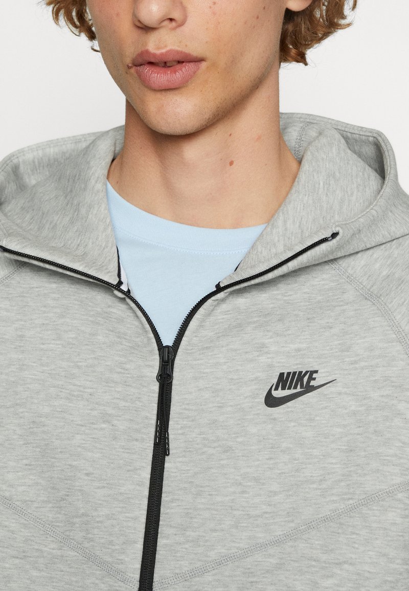 Sudadera con cremallera gris hecha de una tela suave y texturizada. Cuenta con un logo negro de Nike y está superpuesta sobre una camiseta de color azul claro debajo.