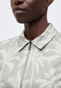 Κοντινό πλάνο ενός ατόμου που φοράει ανοιχτό γκρι πουκάμισο με λευκό μοτίβο φοίνικα και φερμουάρ μπροστά με τράβηγμα σε σχήμα Β.