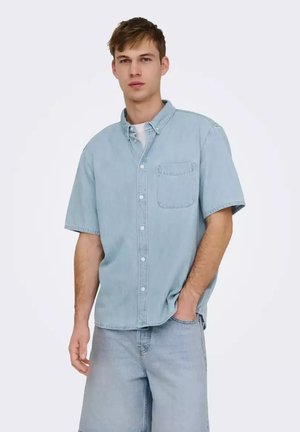 Joven con camisa vaquera azul claro de manga corta y pantalones cortos vaqueros a juego, de pie con una mano en el bolsillo sobre fondo blanco.